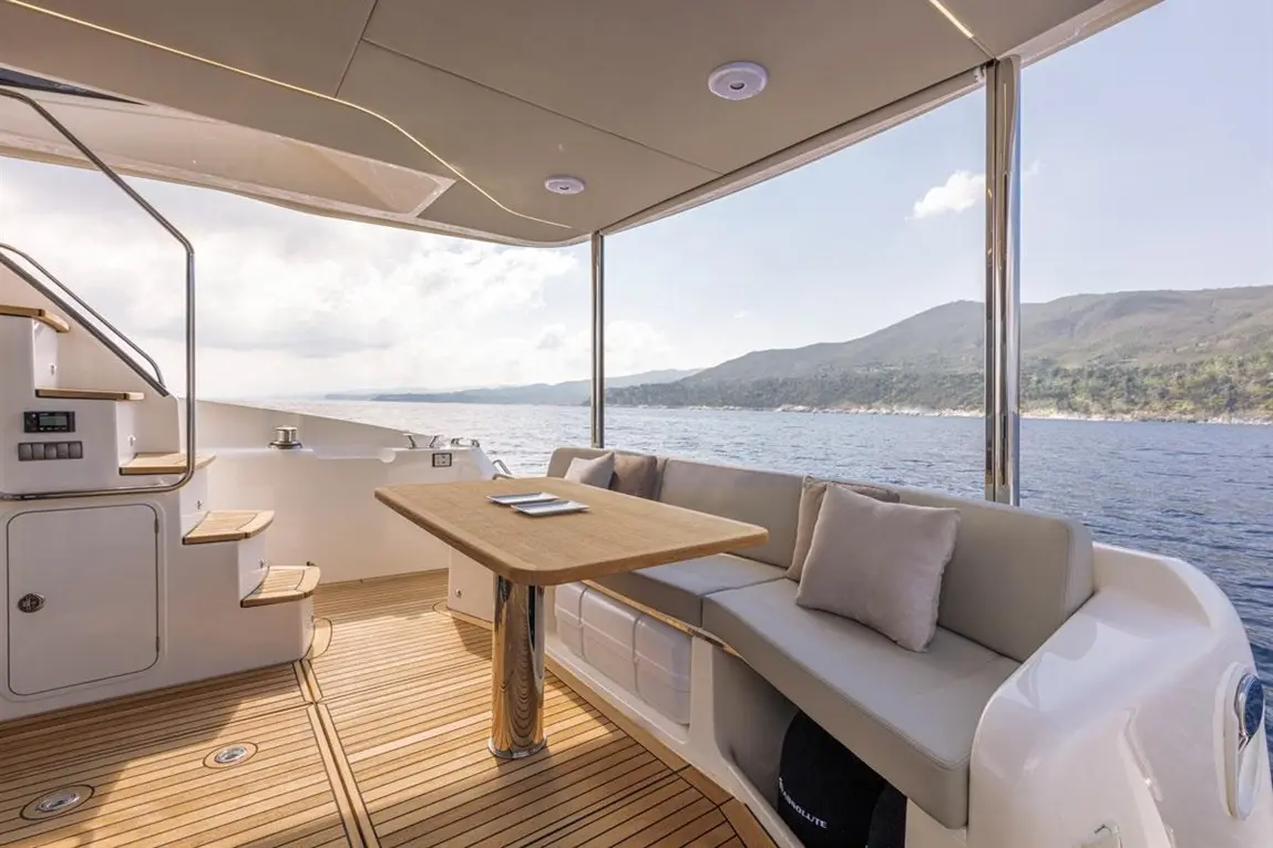 Stylish yacht interior with a wooden table, seating area, and panoramic ocean view against a mountainous backdrop.
